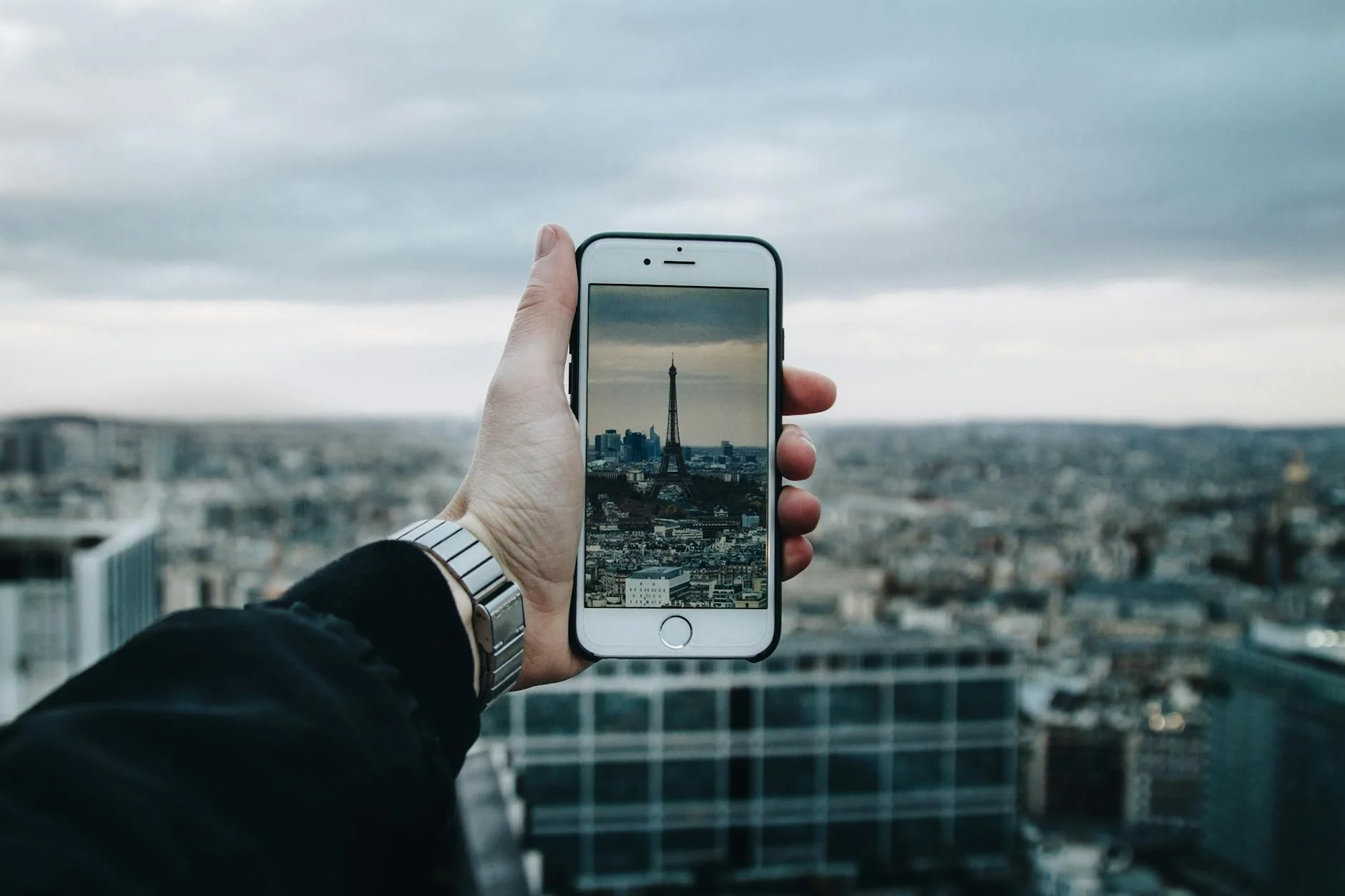 A photorealistic image of a traveler interacting with a local artisan at a vibrant, open-air market, showcasing handmade sustainable products. The scene is bathed in warm, natural light, with sharp focus on the interaction and products, depth of field, 8K hyper-detailed. A person uses a smartphone to capture the Eiffel Tower amidst a panoramic cityscape. — Foto: Valentin Antonucci / Pexels