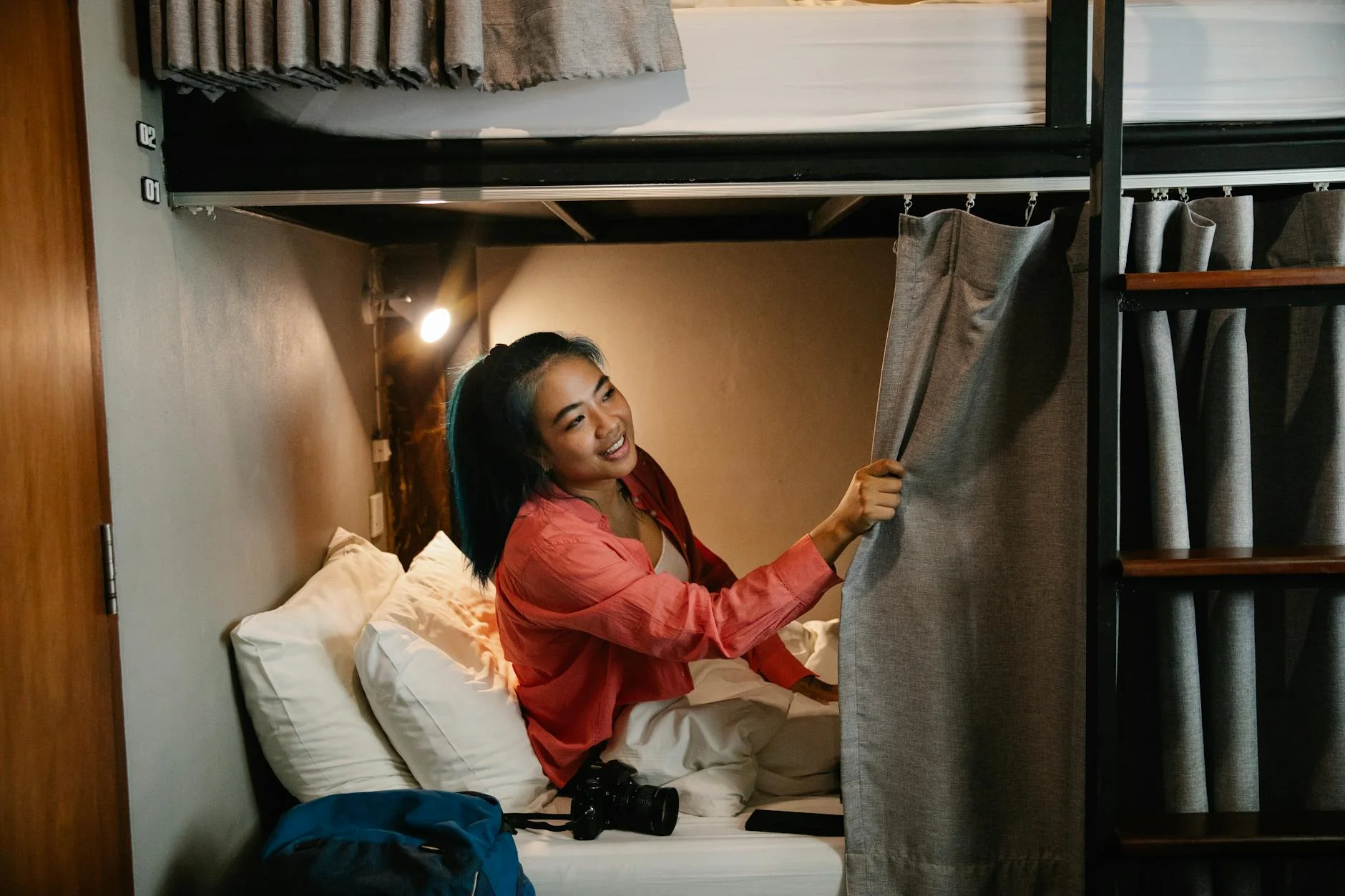 A photorealistic image of a traveler meticulously researching hostels on a laptop, surrounded by maps and travel guides. The screen displays a booking website with filters for 'eco-friendly' and 'budget'. The traveler looks focused and empowered. Professional photography, 8K, cinematic lighting, sharp focus on the laptop screen, depth of field blurring the background, shot on a high-end DSLR. Side view of young smiling ethnic female resting in bed under white blanket with camera and phone and looking away holding curtain in train — Foto: George Pak / Pexels